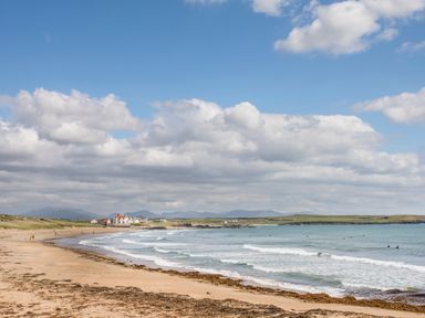 A sandy beach with seaweed and waves with houses and hills in the background at Plas Newydd in Llanfaelog near Rhosneigr