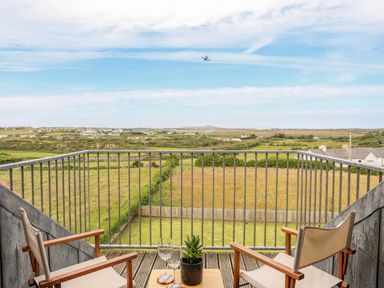 A balcony with two chairs a small table with two glasses and a plant overlooking fields and distant houses at Plas Newydd in Llanfaelog near Rhosneigr