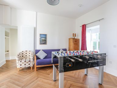 A game room with a foosball table, a blue sofa, an armchair, a wooden cabinet, and a window with red curtains at Plas Newydd in Llanfaelog near Rhosneigr