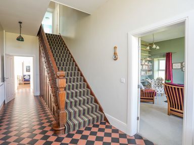 A hallway with checkered floor tiles and a striped carpeted wooden staircase leading upstairs with an adjacent room containing chairs and bookshelves at Plas Newydd in Llanfaelog near Rhosneigr