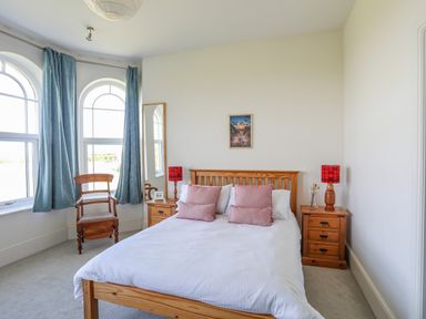 A bedroom with a wooden bed with pink cushions two bedside tables with red lamps a wooden chair and two arched windows with blue curtains at Plas Newydd in Llanfaelog near Rhosneigr
