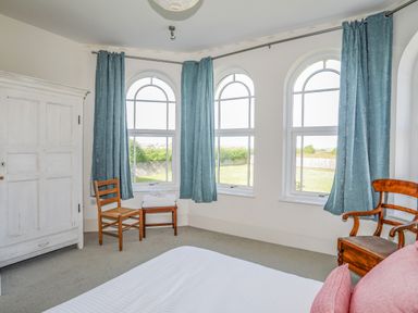 A bedroom with three arched windows with blue curtains two wooden chairs a white wardrobe and a bed with pink pillows at Plas Newydd in Llanfaelog near Rhosneigr