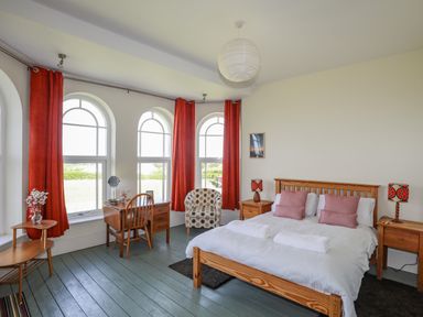 A bedroom with a wooden bed desk chair and polka dot armchair by windows with red curtains at Plas Newydd in Llanfaelog near Rhosneigr
