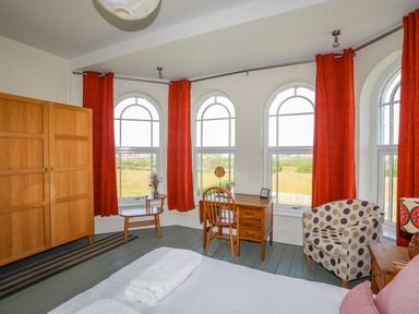 A bedroom with arched windows red curtains a wooden wardrobe a wooden desk and chair a polka dot armchair and a bed with white linens at Plas Newydd in Llanfaelog near Rhosneigr