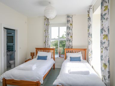 A bedroom with two single beds with white bedding and blue cushions a window with floral curtains and a small bathroom visible through an open door at Plas Newydd in Llanfaelog near Rhosneigr