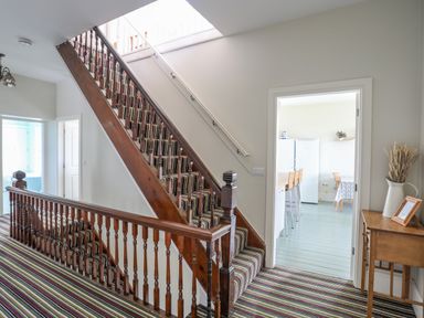 A staircase with striped carpet and wooden railing next to a hallway and door leading to a kitchen at Plas Newydd in Llanfaelog near Rhosneigr