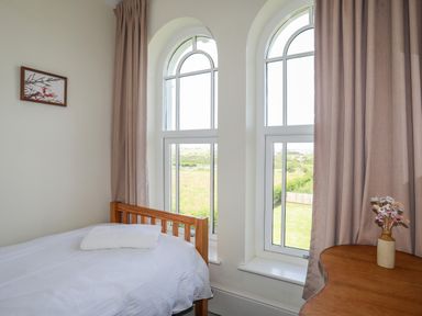 A bedroom with a single bed a folded towel on it two arched windows beige curtains a wooden table and a vase with flowers at Plas Newydd Llanfaelog near Rhosneigr