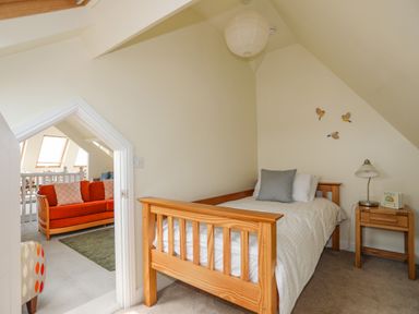 A small bedroom with a single wooden bed a gray pillow bedside table with a lamp and book and a doorway showing a room with an orange sofa at Plas Newydd Llanfaelog near Rhosneigr