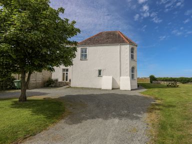 A white two-story house with a red tiled roof next to a tree and a picnic table on a grassy area at Plas Newydd Llanfaelog near Rhosneigr