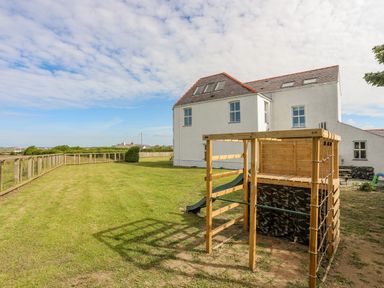 A fenced backyard with a wooden play structure and a white house in the background at Plas Newydd in Llanfaelog near Rhosneigr
