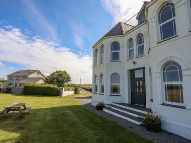 An exterior view of a white house with arched windows and a picnic table on the lawn at Plas Newydd in Llanfaelog near Rhosneigr