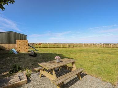 An outdoor area with a wooden picnic table and potted plant a playground slide and climbing frame a lawn and a wire fence at Plas Newydd in Llanfaelog near Rhosneigr