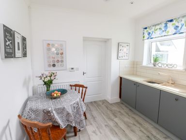 A kitchen with a round table covered with a patterned tablecloth two wooden chairs a bowl of apples flowers on the table grey cabinets and a window with a floral blind at Garth in Pen-y-bryn near Cardigan