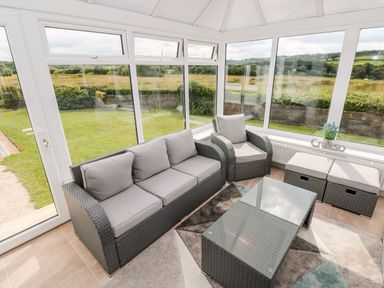 A sunroom with wicker sofa and chair a glass coffee table and ottomans overlooking a grassy outdoor area at Garth Pen-y-bryn near Cardigan