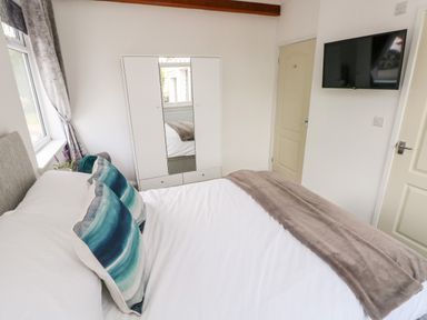A bedroom with a bed with white sheets and blue pillows a white wardrobe with a mirror a wall-mounted television and a window with curtains at Garth in Pen-y-bryn near Cardigan