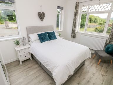 A bedroom with a bed white bedding two blue cushions a bedside table with lamp and plant a gray chair with blue cushion and windows with curtains at Garth in Pen-y-bryn near Cardigan