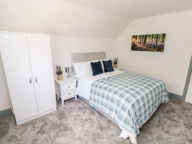 A bedroom with a double bed with blue checkered blanket and two navy pillows two white nightstands with lamps and plants a white wardrobe and a forest picture on the wall at Garth in Pen-y-bryn near Cardigan