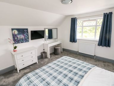 A bedroom with a bed covered in a checkered blanket a white dresser a wall-mounted TV a vanity table with a mirror and stool and a window with blue curtains at Garth in Pen-y-bryn near Cardigan