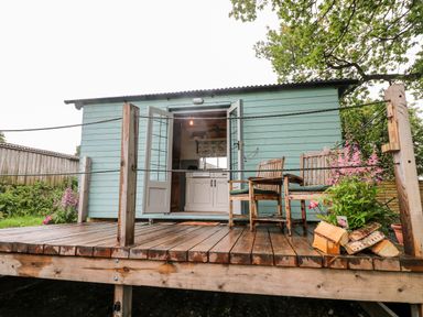 A wooden deck with two chairs and a flower pot outside a light blue cabin at The Lazy Mare by The Water in Admaston near Abbots Bromley