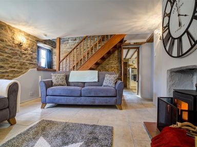 A living room with a sofa clock wood stove staircase and stone walls at Hollytree Cottage in Hook Norton