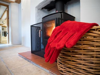 A wood burning stove with fire guard a wicker basket and red gloves at Hollytree Cottage in Hook Norton