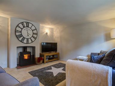 A living room with a fireplace a large wall clock a television on a wooden stand a basket with blankets and a star patterned rug at Hollytree Cottage in Hook Norton