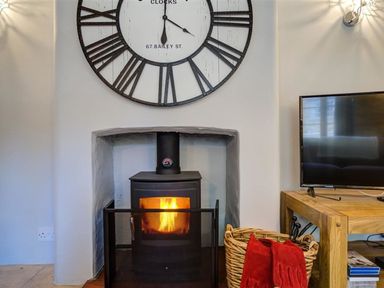 A wood burning stove with a fire inside under a large wall clock next to a basket with red gloves and a wooden table with a television at Hollytree Cottage in Hook Norton