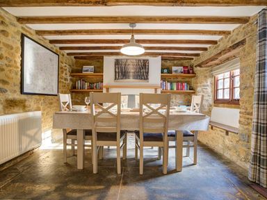 A dining room with a table and six chairs stone walls wooden beams and shelves at Hollytree Cottage in Hook Norton