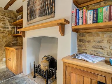 A room with a stone wall wooden shelves and cabinets a fireplace with a large vase and books on shelves at Hollytree Cottage in Hook Norton