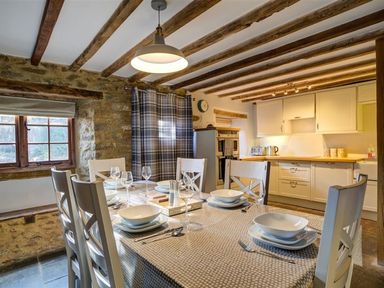 A dining area with a table set for six and a kitchen with cabinets and appliances at Hollytree Cottage in Hook Norton
