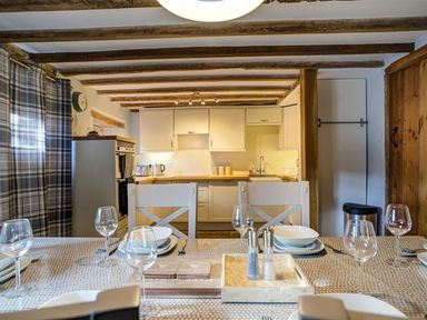 A dining table set with wine glasses and white bowls in front of a kitchen with wooden beams and white cabinets at Hollytree Cottage in Hook Norton