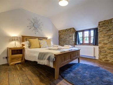 A bedroom with a wooden bed with pillows and towels bedside tables with lamps a round rug and a window with curtains at Hollytree Cottage in Hook Norton