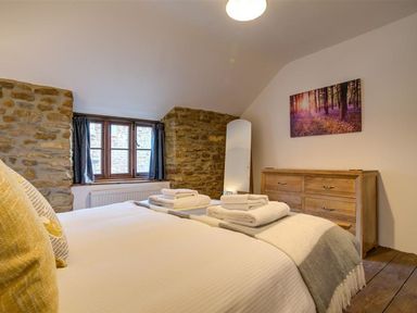 A bedroom with a bed, folded towels, a wooden dresser, a full-length mirror, stone walls, and a window at Hollytree Cottage in Hook Norton