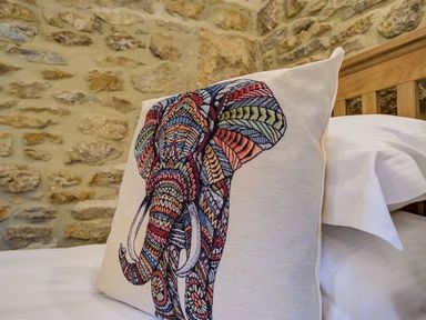 A decorative pillow with a colorful elephant design on a bed with white sheets and a wooden headboard in front of a stone wall at Hollytree Cottage in Hook Norton