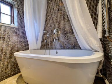 A bathroom with a white bathtub mosaic tiled walls a window and white shower curtains at Hollytree Cottage in Hook Norton