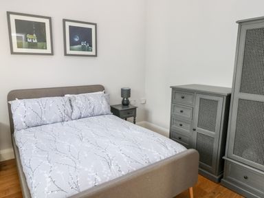 A bedroom with a bed with patterned bedding a bedside table with a lamp two framed pictures on the wall and grey wooden furniture at Ramsey Cottage in Milford Haven