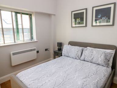 A bedroom with a bed, bedside table with a lamp, two framed pictures on the wall and a window with blinds at Ramsey Cottage in Milford Haven