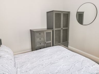 A bedroom with a bed with patterned bedding a grey wardrobe and dresser and a round wall mirror at Ramsey Cottage in Milford Haven
