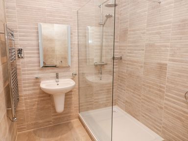 A bathroom with a wall-mounted sink a rectangular mirror a shower area with a glass partition and tiled walls at Ramsey Cottage in Milford Haven