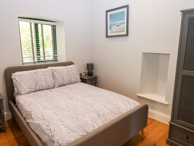 A bedroom with a bed with patterned bedding two bedside tables a lamp a framed picture on the wall a window with blinds and a wooden wardrobe at Ramsey Cottage in Milford Haven