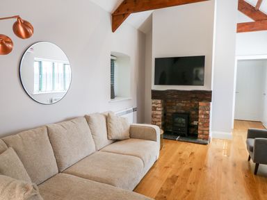 A living room with a beige sofa a round mirror on the wall a wall-mounted TV above a brick fireplace and wooden beams at Ramsey Cottage in Milford Haven