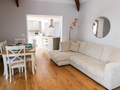 An open living and dining area with a beige sofa a wooden dining table set with plates and bowls and a kitchen with white cabinets and appliances at Ramsey Cottage in Milford Haven