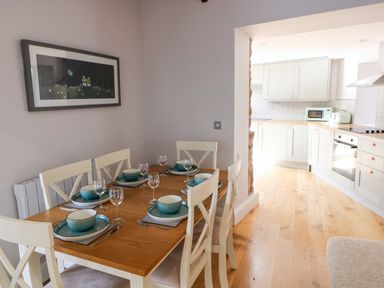 A dining area with a wooden table set with plates, bowls, glasses, and cutlery next to a kitchen with white cabinets and wooden countertops at Ramsey Cottage in Milford Haven