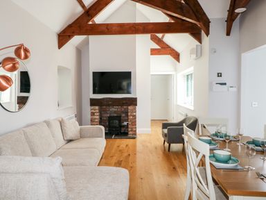 A living room with a beige sofa a wall-mounted TV above a brick fireplace wooden ceiling beams and a dining table set with green dishes at Ramsey Cottage in Milford Haven