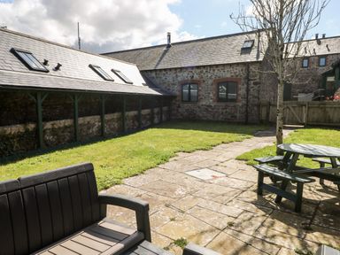 A garden with a stone patio, wooden bench, picnic table, tree, and stone buildings at Ramsey Cottage in Milford Haven