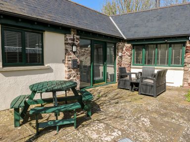 An outdoor patio with a green wooden picnic table and four benches and two gray single chairs with a small table outside a stone and white plaster building with green framed windows and door at Ramsey Cottage in Milford Haven