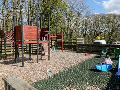 An outdoor playground with wood chip ground cover red and black climbing structures plastic rocker toys and a plastic chair at Ramsey Cottage in Milford Haven