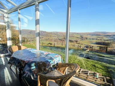 A conservatory with a table covered by a patterned cloth and two wicker chairs with a view of green fields and hills at Ty Twmp Tump Cottage in Tal-Y-Bont-On-Usk