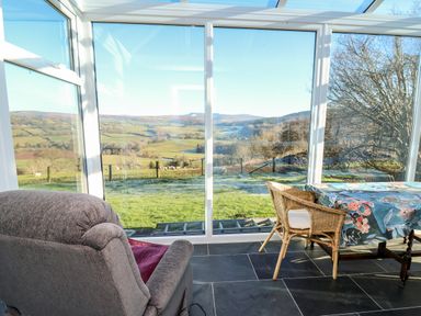 A room with a chair and table covered with a floral cloth with large glass windows showing a countryside view at Ty Twmp Tump Cottage in Tal-Y-Bont-On-Usk