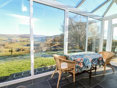 A conservatory with a table covered by a floral tablecloth and two wicker chairs with a view of a green landscape and hills at Ty Twmp Tump Cottage in Tal-Y-Bont-On-Usk